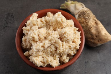 Bowl of tasty prepared horseradish and root on grey table, closeup