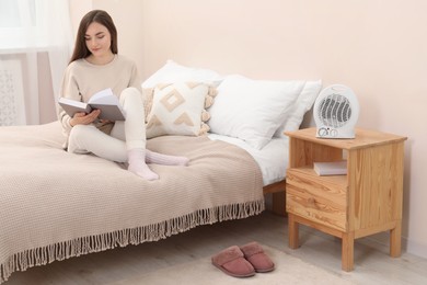 Photo of Woman reading book near portable electric fan heater in bedroom