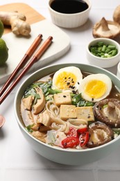 Photo of Noodle soup. Bowl of delicious ramen, ingredients and chopsticks on white tiled table, closeup