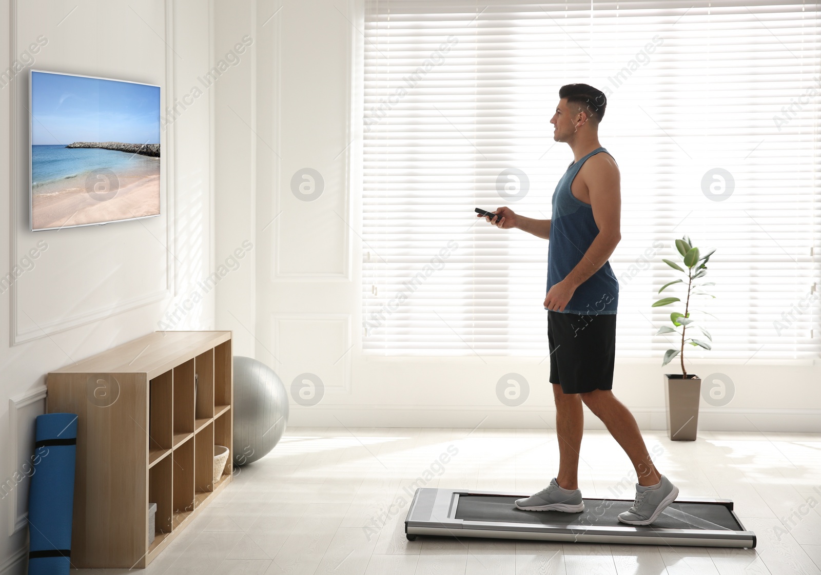 Image of Sporty man with remote control training on walking treadmill and watching TV at home