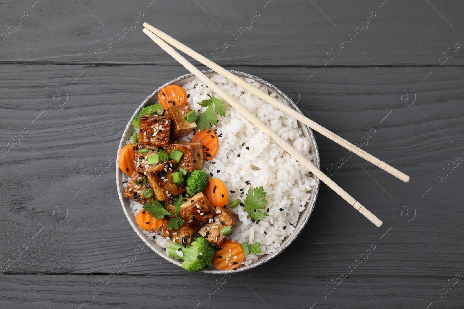 Photo of Bowl of rice with fried tofu, broccoli and carrots on grey wooden table, top view