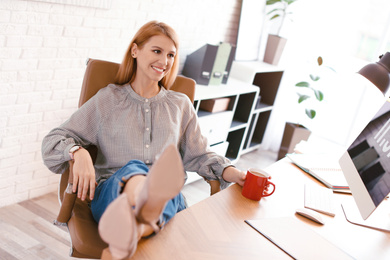 Young woman with cup of drink relaxing at table in office during break