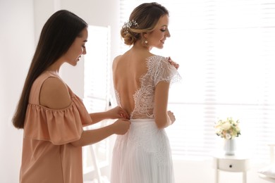 Young woman helping bride to put on wedding dress in room