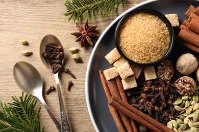 Photo of Plate with different aromatic spices, spoons and fir branches on wooden table, flat lay