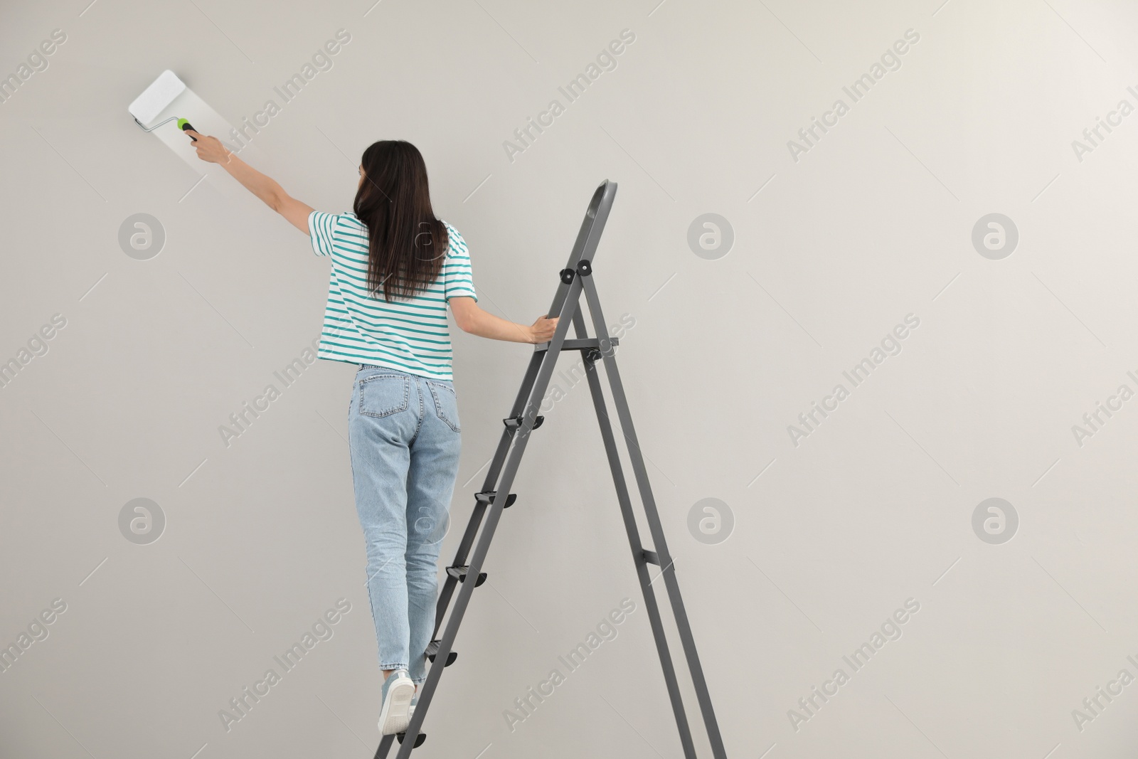 Photo of Young woman painting wall with roller on ladder, back view