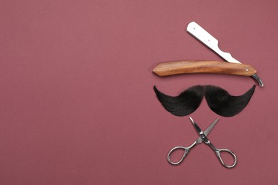 Photo of Artificial moustache and barber tools on burgundy background, flat lay. Space for text