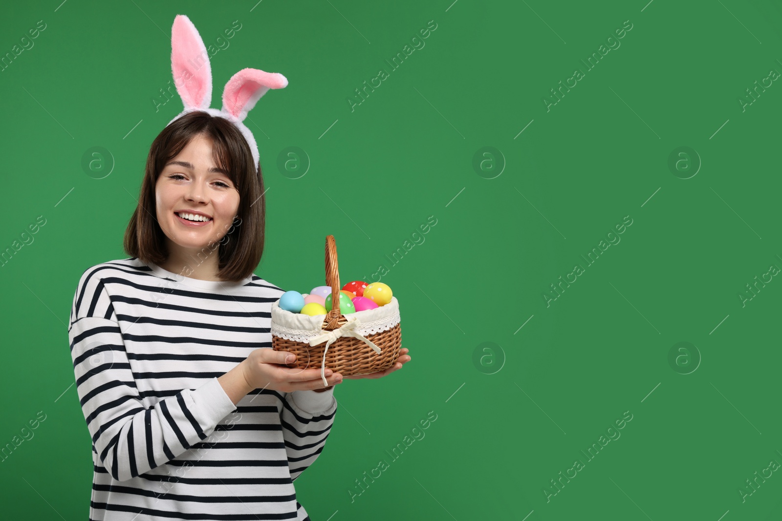 Photo of Easter celebration. Happy woman with bunny ears and wicker basket full of painted eggs on green background, space for text