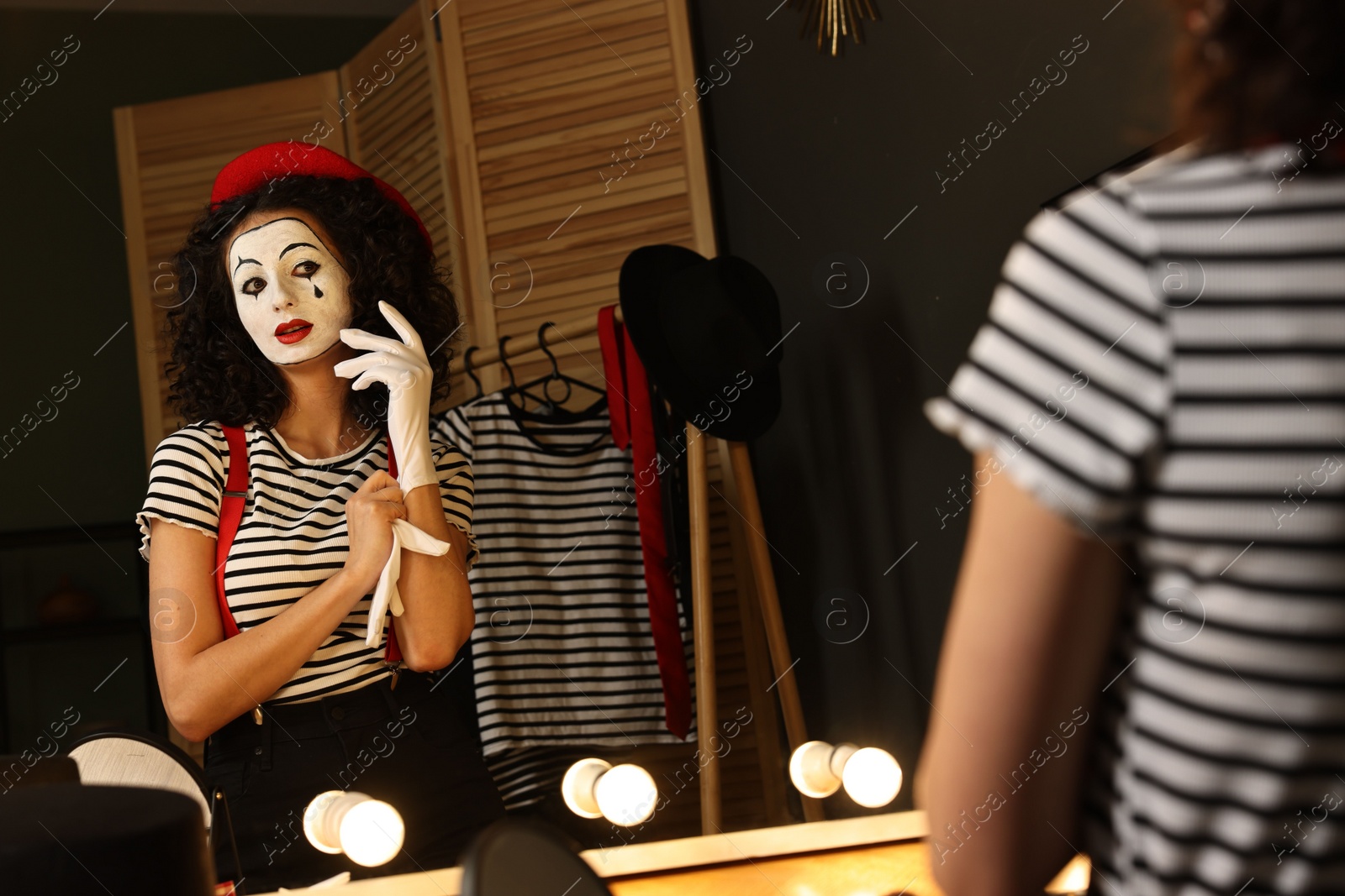 Photo of Young woman in mime costume putting gloves near mirror indoors