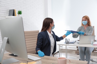 Office employee in mask and gloves giving clipboard to her colleague at workplace
