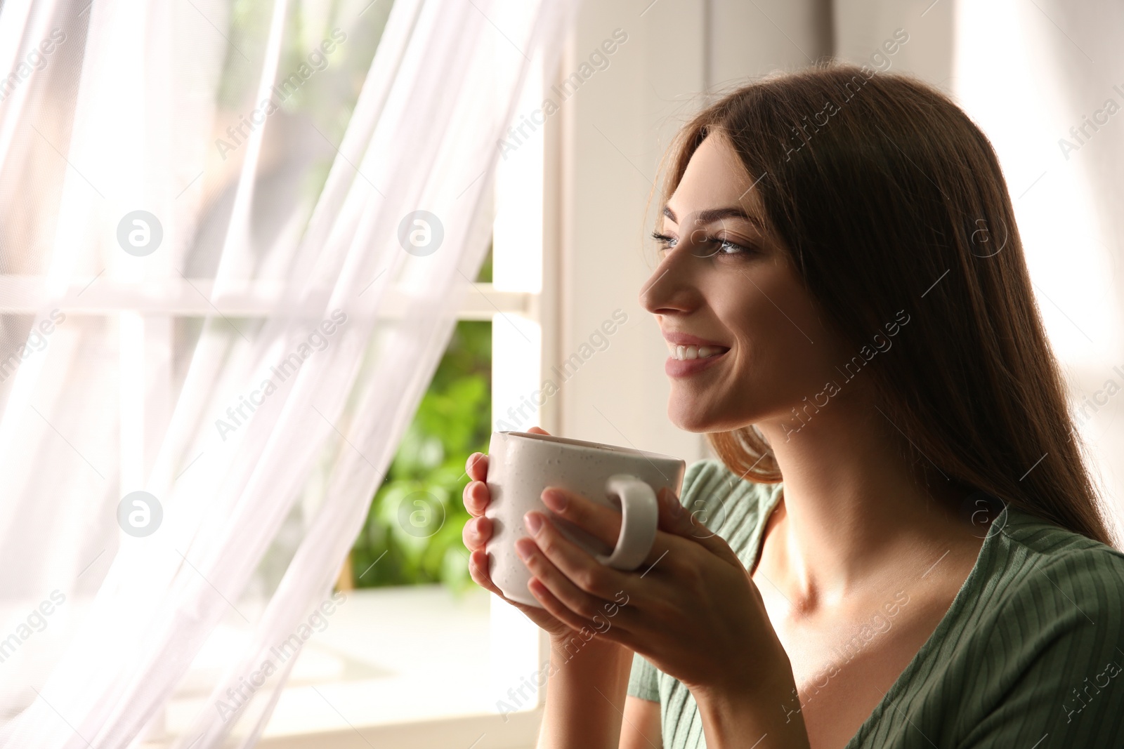 Photo of Beautiful young woman with cup of drink near window at home