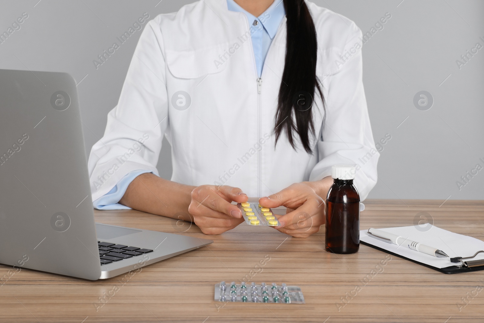 Photo of Professional pharmacist with pills and laptop at table against light grey background, closeup