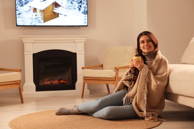 Photo of Young woman with cup of tea relaxing on floor near fireplace at home