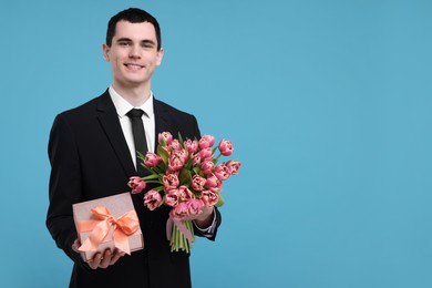 Photo of Happy young man with beautiful bouquet and present on light blue background. Space for text