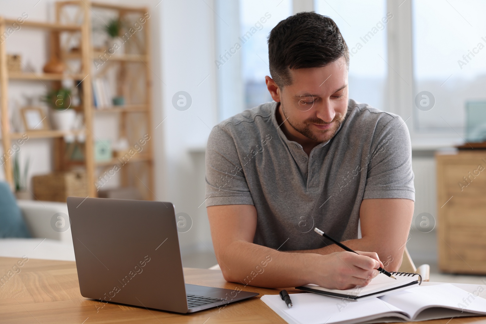 Photo of Man writing notes while working on laptop at wooden desk in room