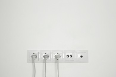 Many power sockets with plugs, ethernet and TV coax plates on white wall indoors. Space for text