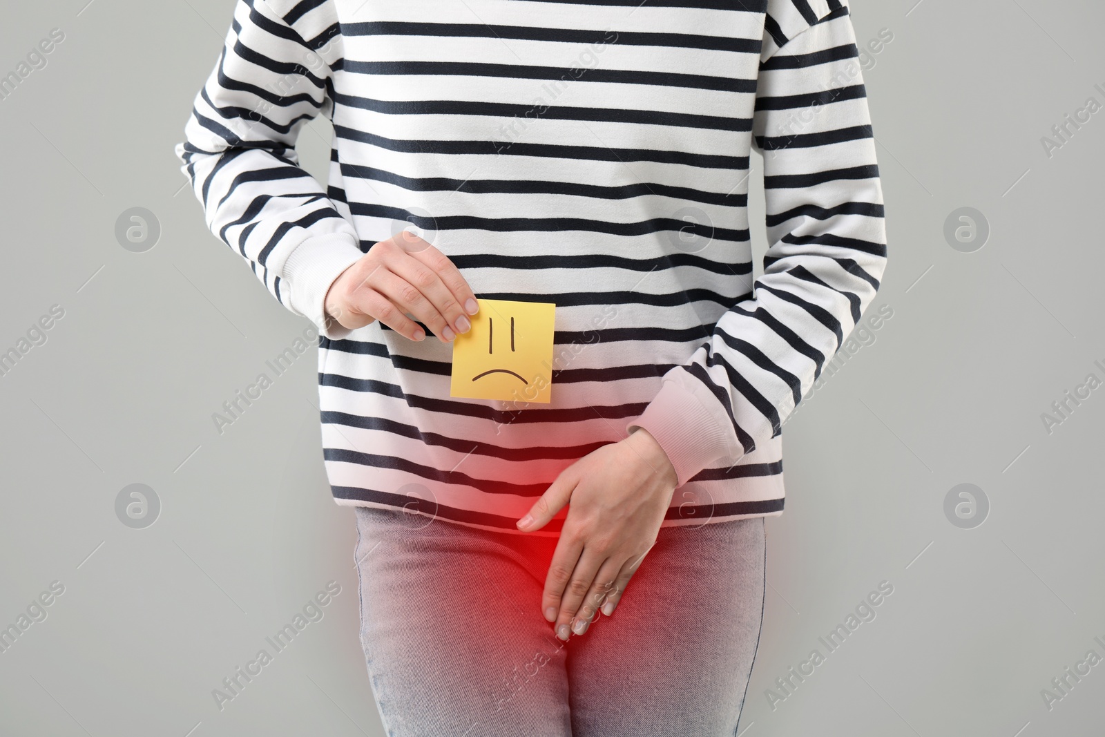 Image of Woman holding sticky note with drawn sad face and suffering from cystitis symptoms on light grey background, closeup