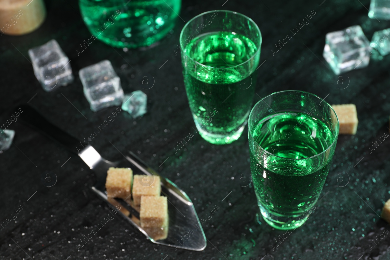 Photo of Absinthe in shot glasses, spoon, brown sugar and ice cubes on gray table, closeup. Alcoholic drink
