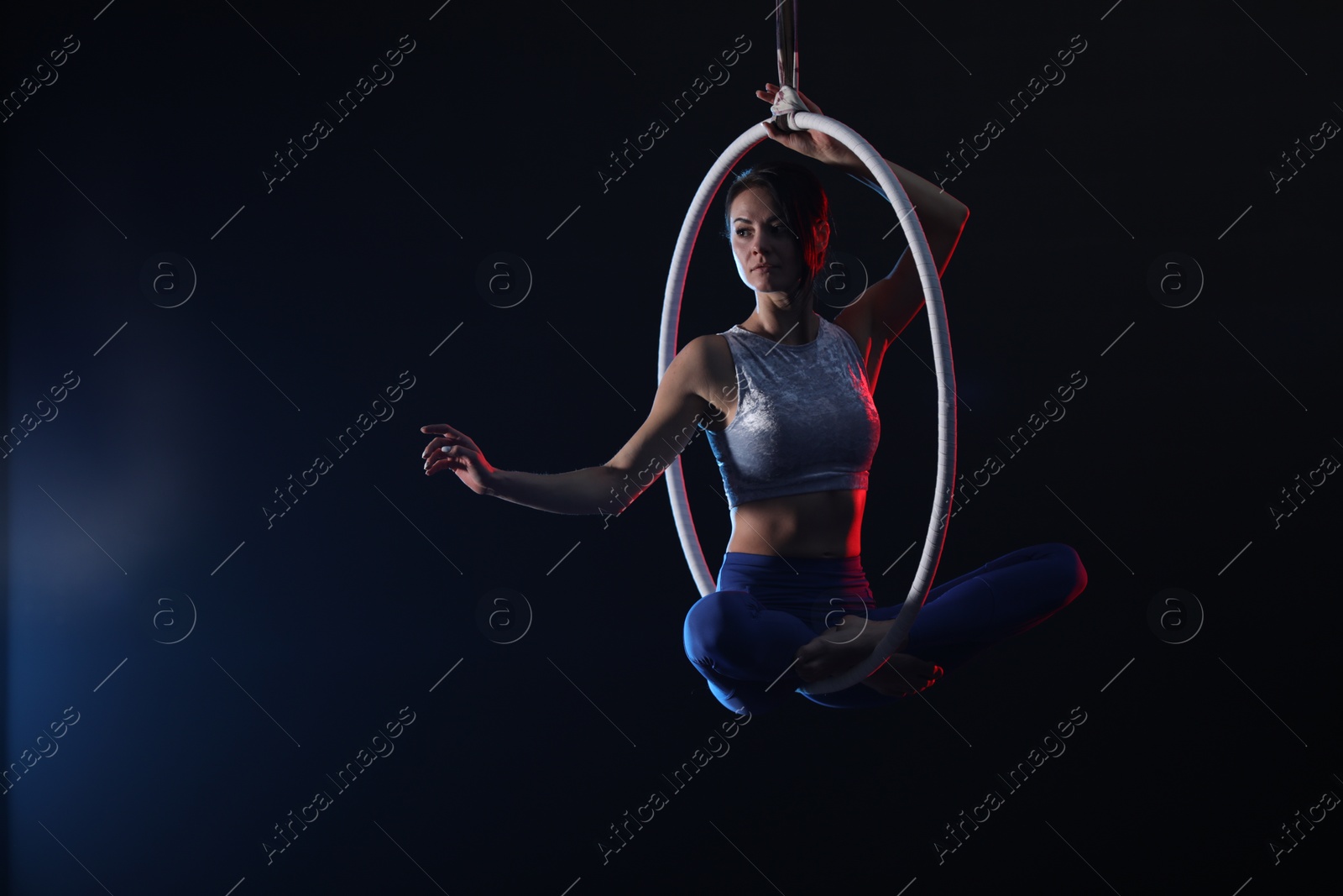 Photo of Young woman performing acrobatic element on aerial ring against dark background