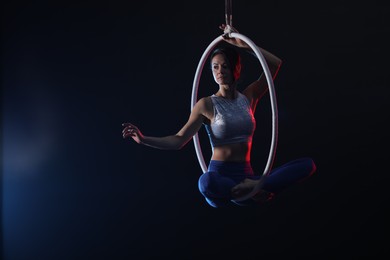 Young woman performing acrobatic element on aerial ring against dark background