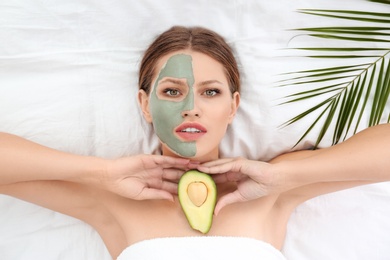 Photo of Beautiful woman holding avocado near her face with clay mask on white fabric, above view