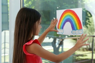 Photo of Little girl with picture of rainbow near window indoors.  Stay at home concept