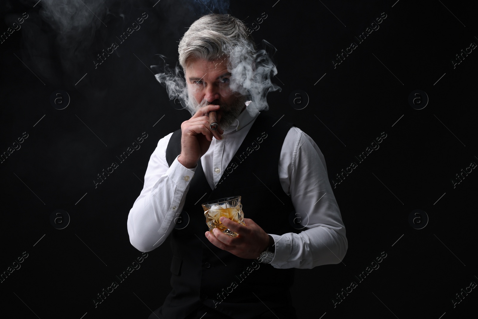 Photo of Bearded man with glass of whiskey smoking cigar against black background