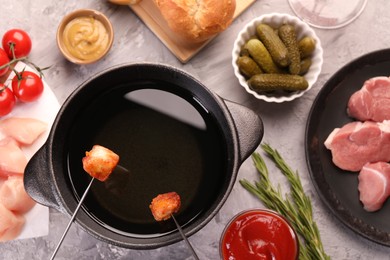 Photo of Fondue pot, forks with fried meat pieces, pickled cucumbers and other products on grey textured table, flat lay