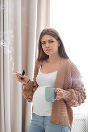 Photo of Young pregnant woman with cup of coffee smoking cigarette at home. Harm to unborn baby