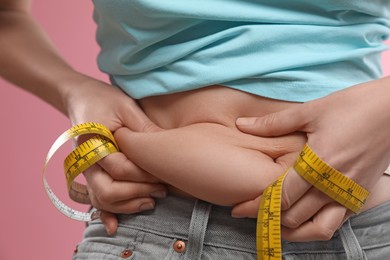 Photo of Woman with measuring tape touching belly fat on pink background, closeup. Overweight problem