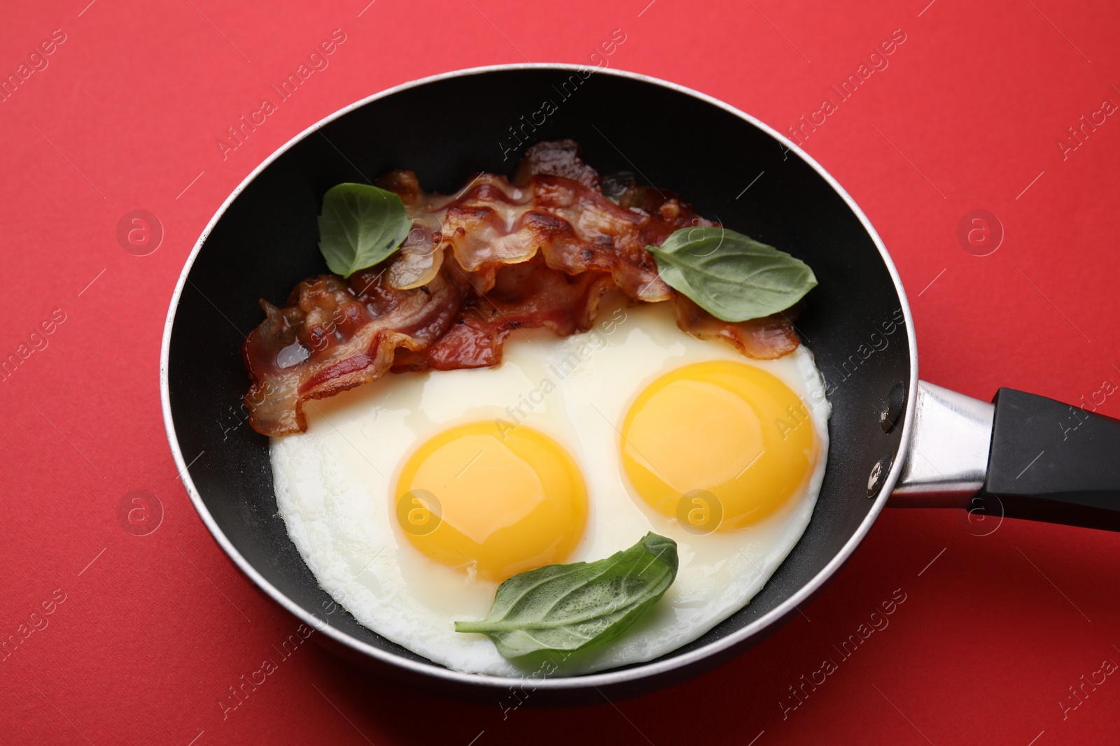 Photo of Fried eggs, bacon and basil in frying pan on red background
