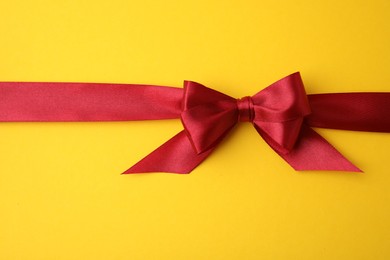 Red satin ribbon with bow on yellow background, top view