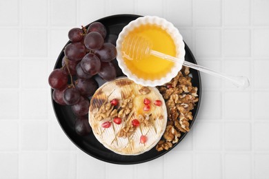 Plate with tasty baked camembert, honey, grapes, walnuts and pomegranate seeds on white tiled table, top view