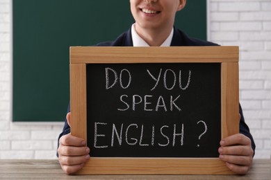 Photo of Teacher holding small chalkboard with inscription Do You Speak English? at table in classroom, closeup