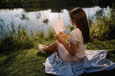 Young woman reading book near lake on sunny day