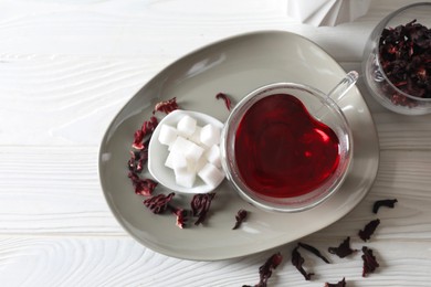 Photo of Delicious hibiscus tea in cup, sugar cubes and dry roselle petals on white wooden table, top view. Space for text