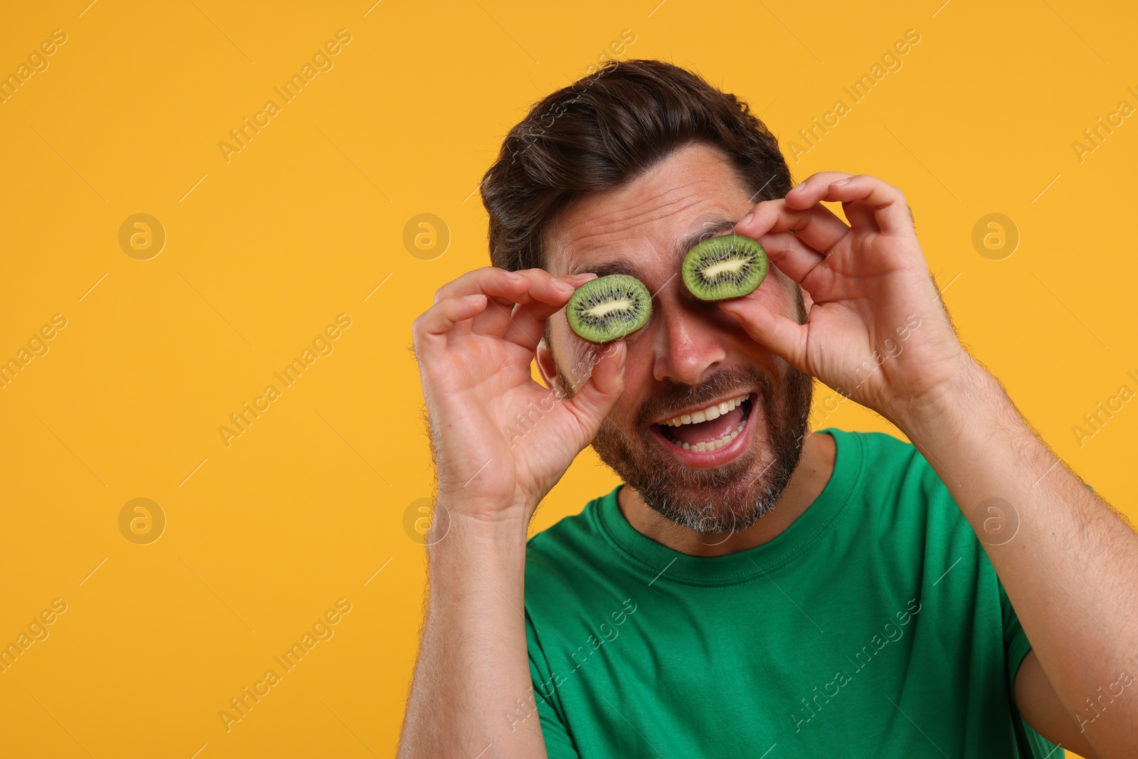 Photo of Emotional man holding halves of kiwi near his eyes on orange background, space for text