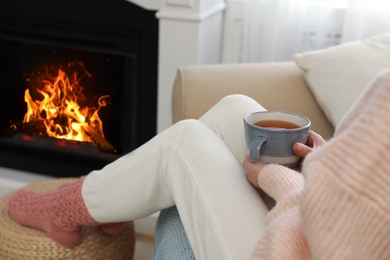 Photo of Woman with tea sitting near fireplace at home, closeup