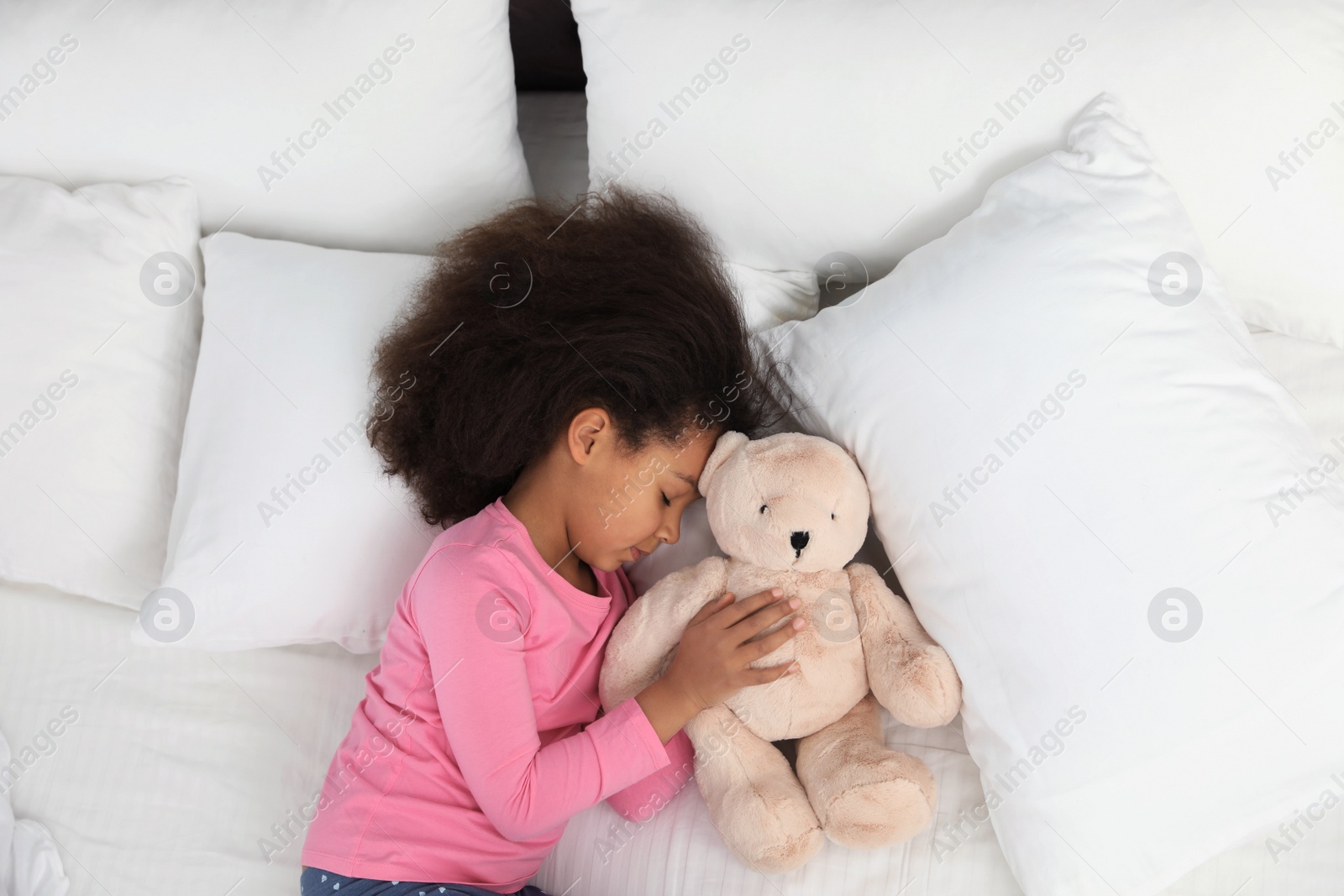 Photo of Cute little African-American girl with teddy bear sleeping in bed, top view