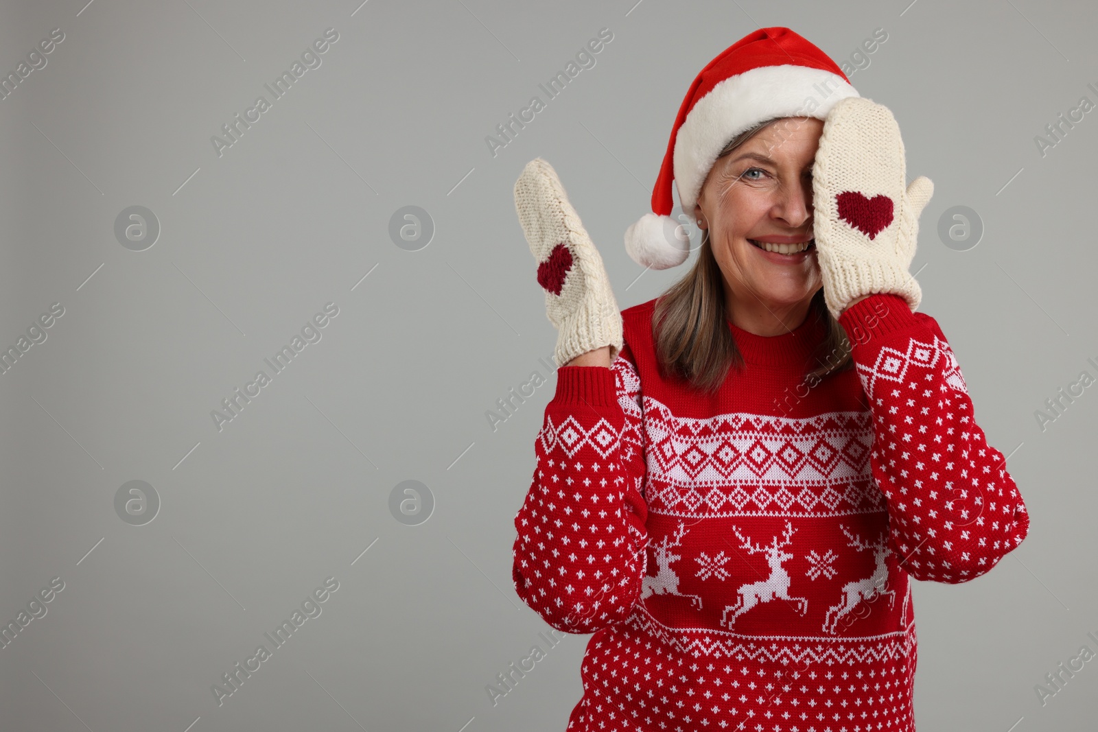 Photo of Happy senior woman in Christmas sweater, Santa hat and knitted mittens on grey background. Space for text