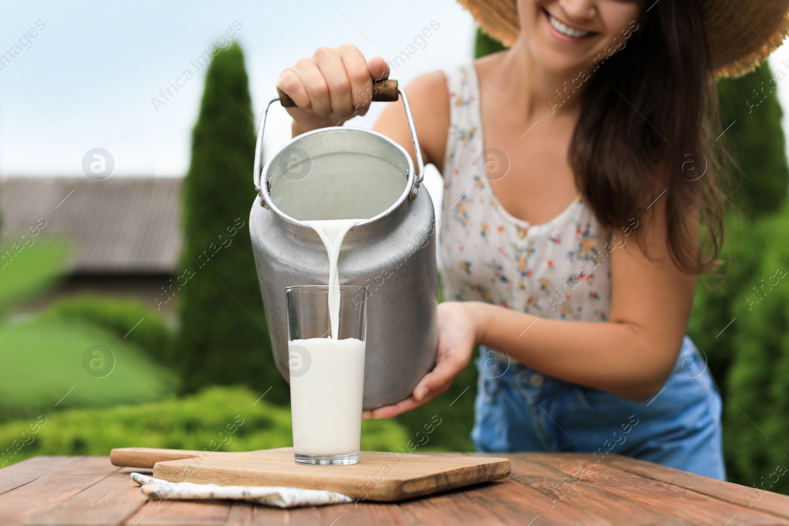 Photo of Smiling woman pouring fresh milk from can into glass at wooden table outdoors, closeup