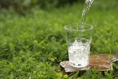 Photo of Pouring fresh water into glass on stone in green grass outdoors. Space for text