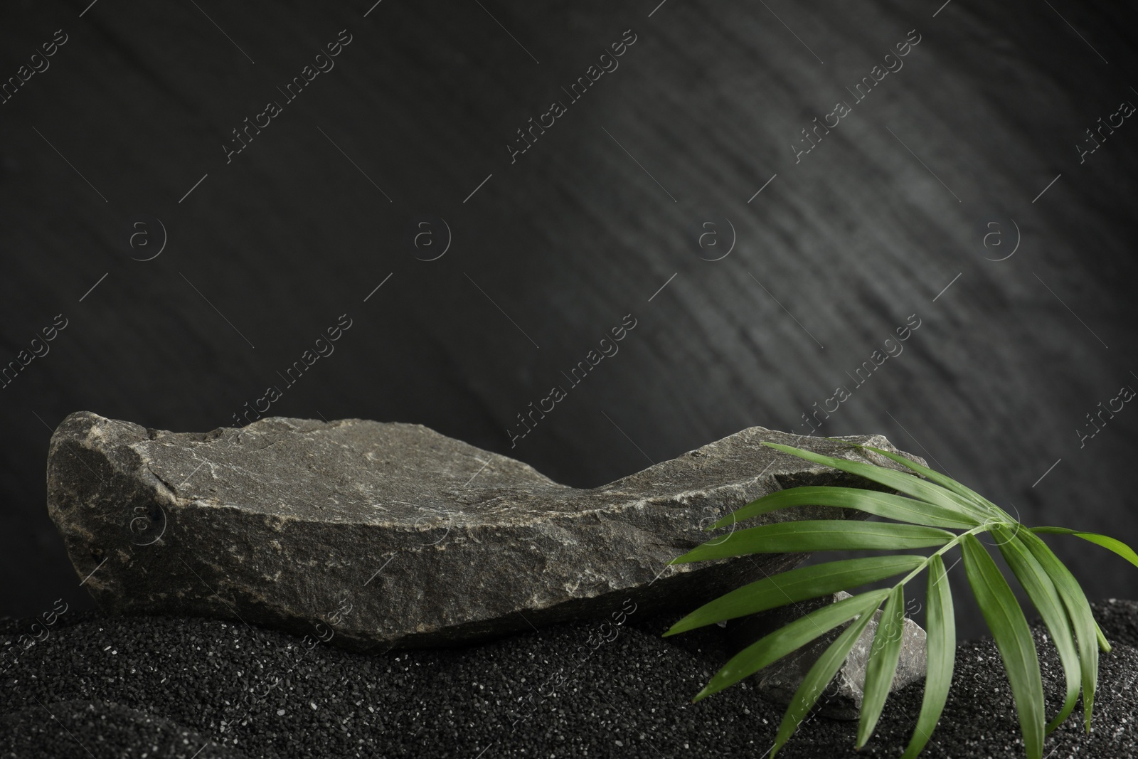 Photo of Presentation of product. Stone podium and palm leaves on black sand against dark textured background. Space for text