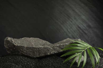 Photo of Presentation of product. Stone podium and palm leaves on black sand against dark textured background. Space for text