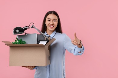Happy unemployed woman with box of personal office belongings showing thumbs up on pink background, space for text
