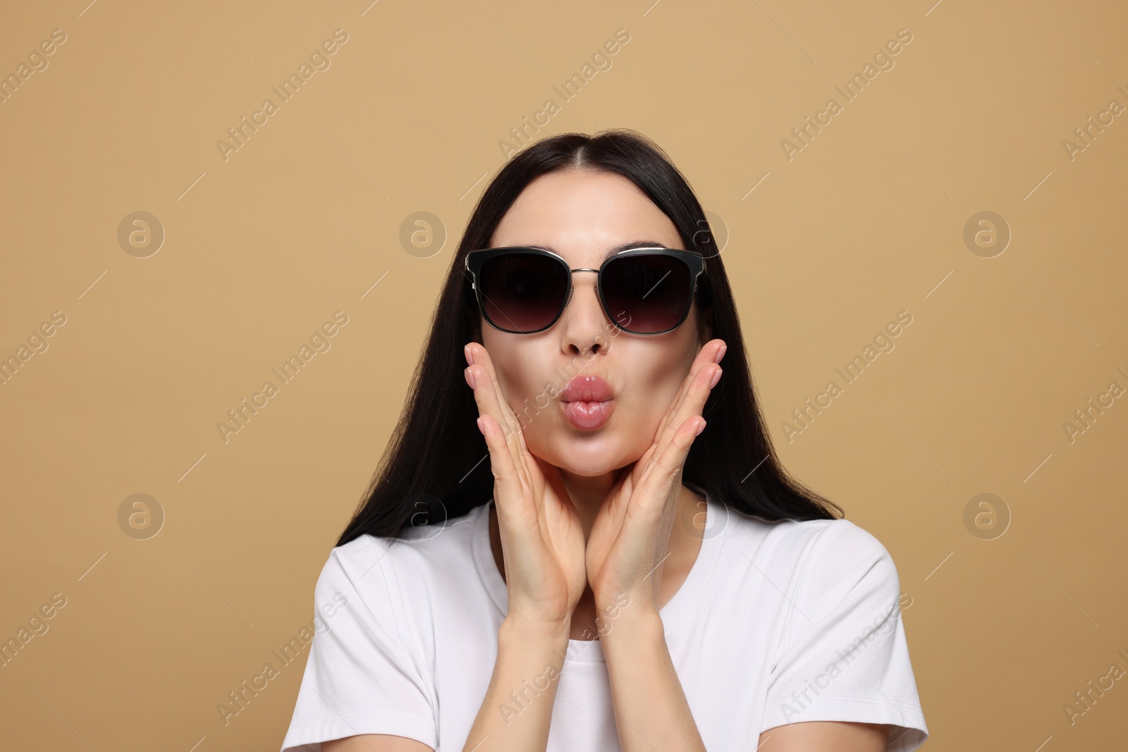 Photo of Beautiful young woman in stylish sunglasses blowing kiss on beige background