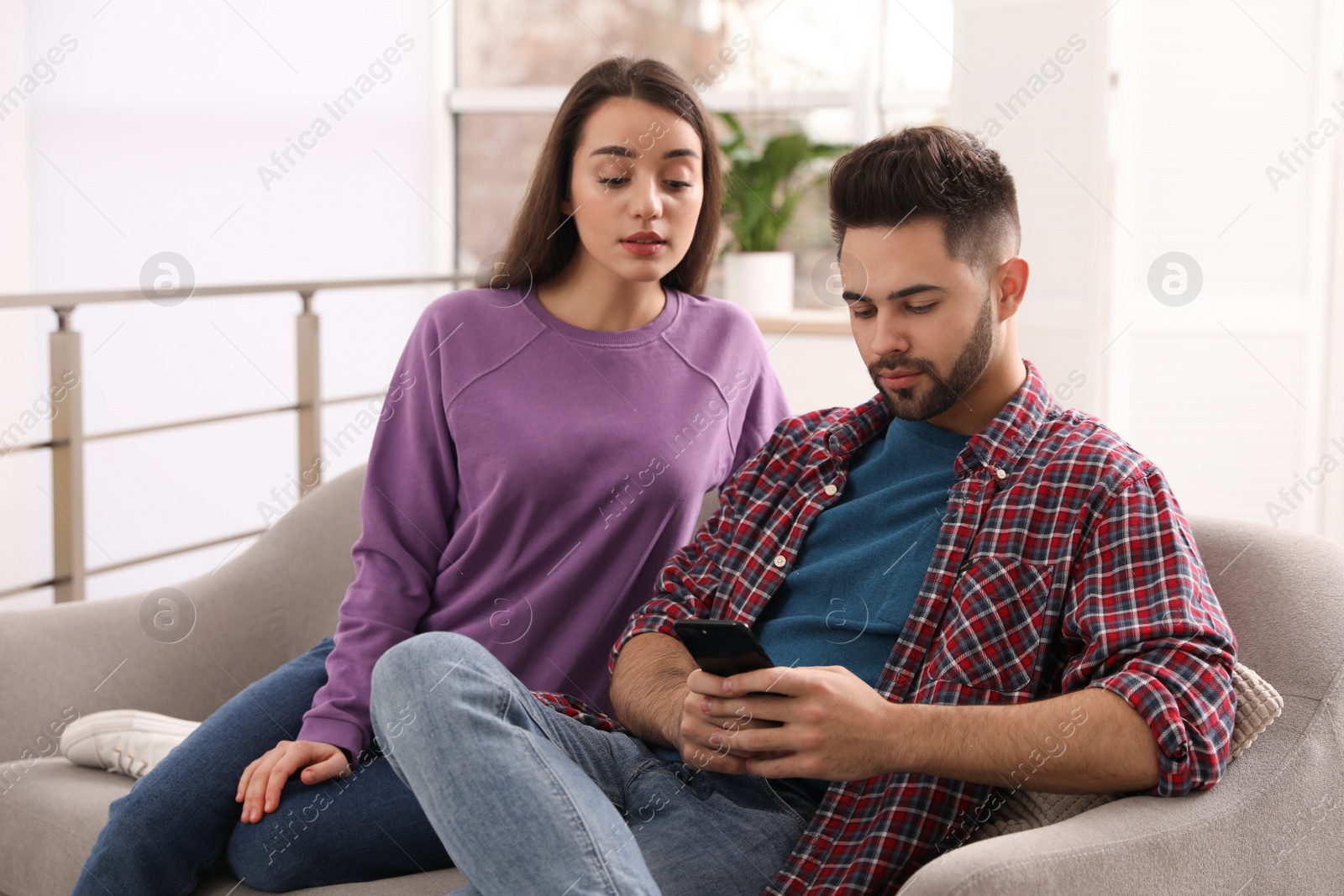 Photo of Distrustful young woman peering into boyfriend's smartphone at home