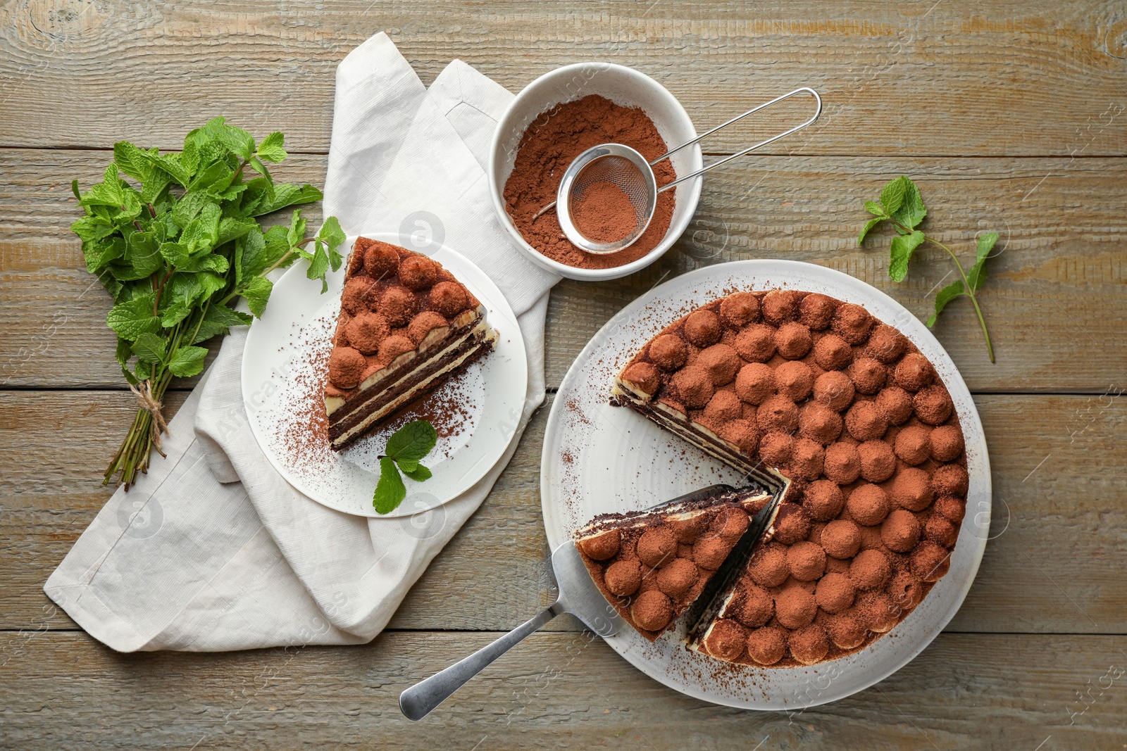 Photo of Delicious tiramisu cake, cocoa powder, server and mint leaves on wooden table, flat lay