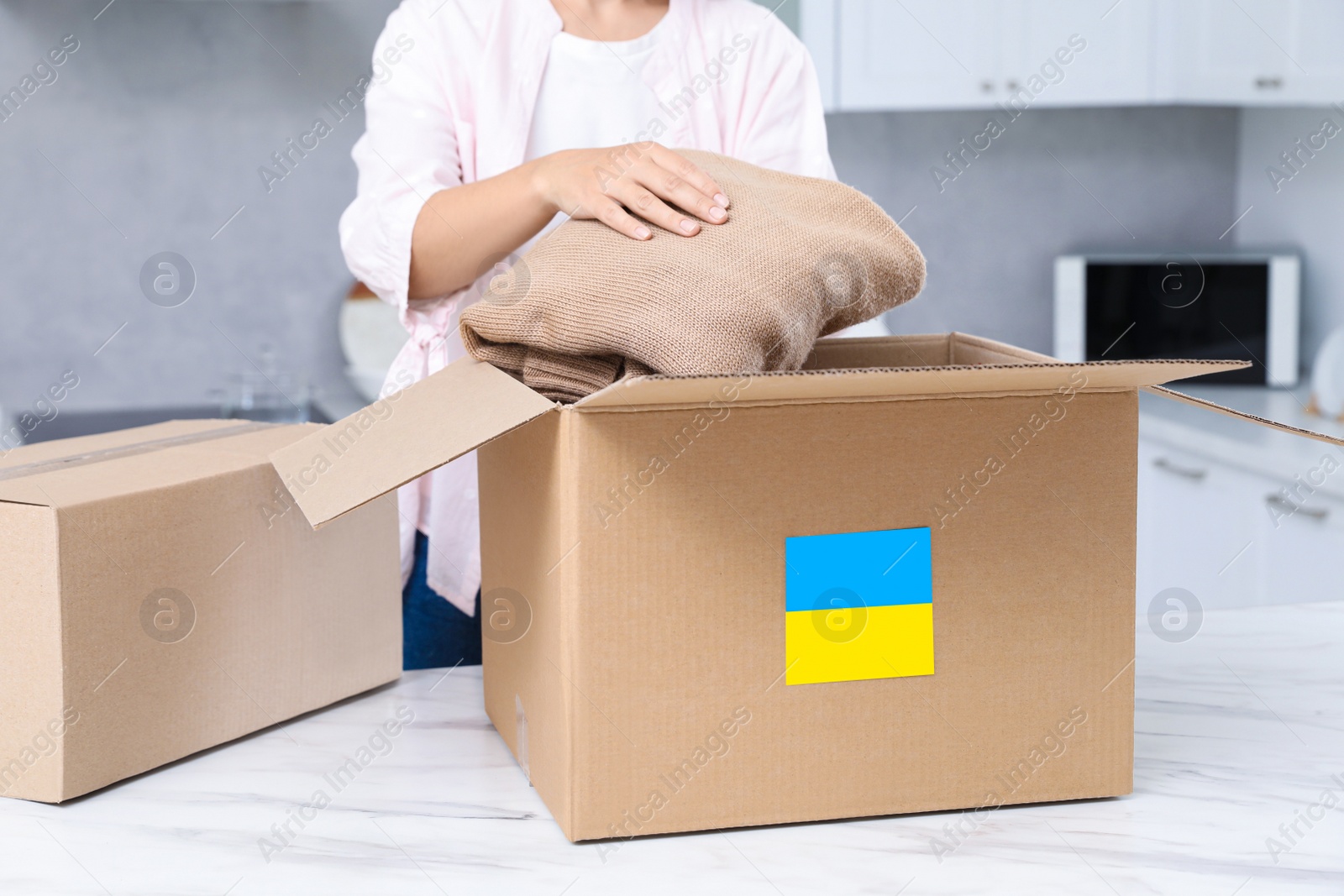 Photo of Humanitarian aid for Ukraine. Woman putting clothes in donation box at white table indoors, closeup