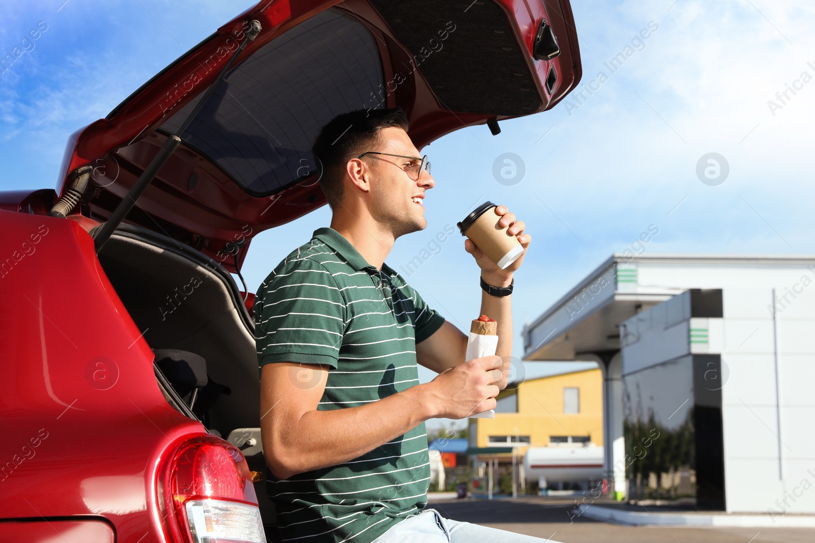 Photo of Young man with hot dog drinking coffee near car at gas station