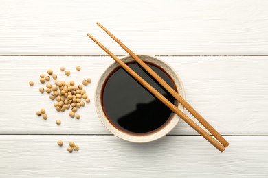 Photo of Tasty soy sauce in bowl, chopsticks and soybeans on white wooden table, top view